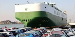 A large green and white Wallenius Wilhelmsen ship docked at a port with many cars parked nearby.