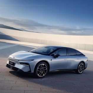 A silver Xpeng P7+ electric car parked on a paved area with a clear blue sky and distant mountains in the background.