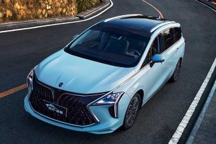 A light blue MPV drives on a coastal winding road, with lush greenery and the sea in the background, highlighting its sleek design and scenic appeal.