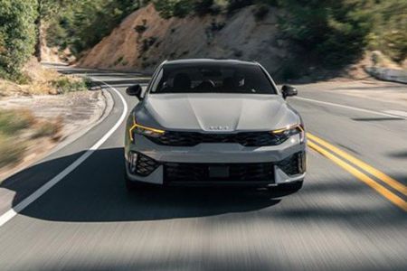 A gray Kia sedan driving on a winding mountain road, with lush greenery and rocky terrain in the background, highlighting its dynamic design and performance.