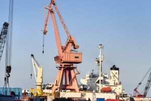 A large red crane and a ship at a busy port under a clear blue sky.