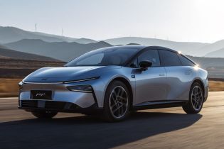 A silver Xpeng P7+ electric car driving on a road with mountains and wind turbines in the background.