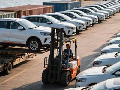 A forklift handling a car at a port with rows of white cars and ships in the background.