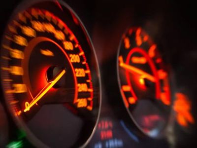 Close-up of a car's illuminated dashboard, featuring a glowing speedometer and tachometer with red/orange backlighting, capturing the thrill of high-speed driving.