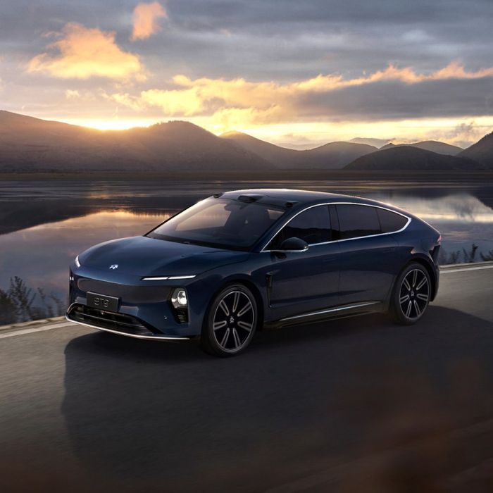 A dark blue NIO ET9 electric car driving by a lake at sunset with mountains in the background.