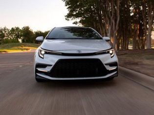 White Toyota sedan driving on road: front view, sleek grille, tree-lined backdrop in soft light.