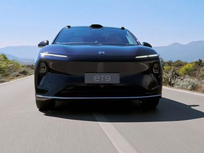 Dark NIO ET9 electric car: front view, driving on road with mountain/vegetation backdrop under blue sky.