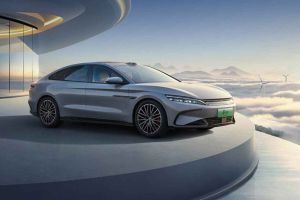 A silver BYD electric sedan displayed on a platform with a futuristic building and cloudscape background.