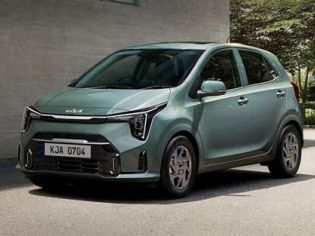 A light green Kia hatchback, with license plate KJA 0704, is parked on a paved area beside a concrete building and greenery, showcasing its modern and compact design.