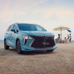 A light blue MPV is parked on a sandy beach, with the sea and a family enjoying the shore in the background, highlighting its stylish design and family-friendly appeal.