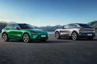 Two Xiaomi electric cars, one green and one silver, parked on a paved area with a scenic backdrop of mountains and a lake.