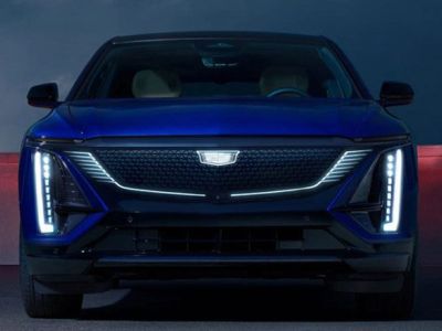 Front view of a blue Cadillac electric vehicle, featuring futuristic LED headlights and a distinctive grille, set against a dramatic nighttime backdrop.