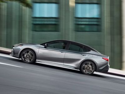 Side profile shot of sleek silver sport sedan driving on urban road, showcasing sculpted body lines, alloy wheels and modern fastback rear styling