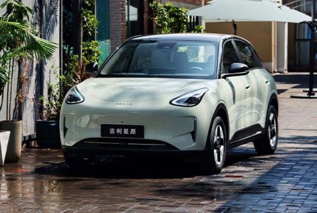 A light green Geely Xingyuan electric car parked on a wet brick road with plants and umbrellas in the background.