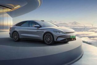 A silver BYD electric sedan displayed on a platform with a futuristic building and cloudscape background.