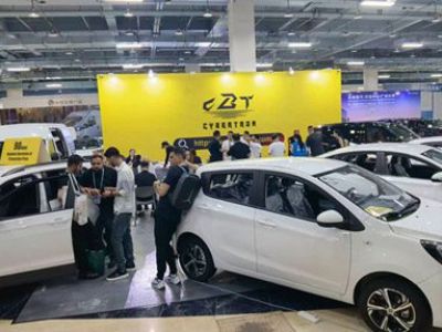A car exhibition scene with several white cars on display, a large yellow "CBT CYBERTRON" backdrop, and people interacting, set in an indoor exhibition hall.