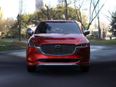 Front view of a red Mazda SUV, featuring its distinctive grille and Mazda logo, parked on a road in a park-like urban setting with trees and buildings in the background.