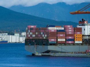 A large container ship loaded with colorful containers docked at a port, with mountains in the background.