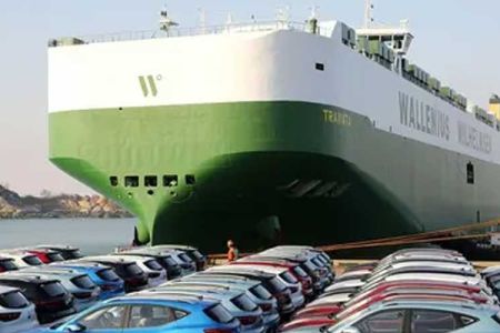 A large green and white Wallenius Wilhelmsen ship docked at a port with many cars parked nearby.