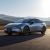 A silver Xpeng P7+ electric car driving on a road with mountains and wind turbines in the background.