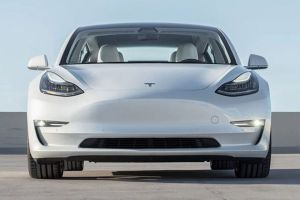 Front view of a white Tesla Model 3 sedan, featuring sleek headlights, a minimalist front design, and a spacious interior, parked on a concrete surface under a clear sky.