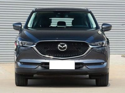 Front view of a dark gray Mazda SUV, parked in front of a corrugated metal wall, featuring its grille, headlights, and sleek front design with a blank license plate.