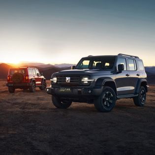 Two off - road SUVs (one gray, one red) parked on an open plain with a sunset and mountainous background.