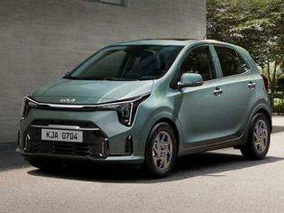 A light green Kia car with the license plate "KJA 0704" parked beside a gray building in a sunny outdoor setting.