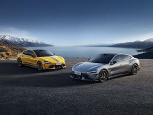 Two sports cars, one yellow with gray stripes and one silver, parked on a paved area with a lake and snow-capped mountains in the background, highlighting their sleek designs and scenic setting.