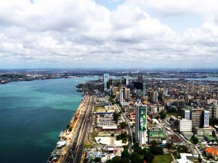 Aerial view of a coastal city with tall buildings, a busy road along the waterfront, and a large body of water under a partly cloudy sky.