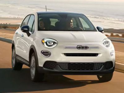 A white Fiat compact SUV drives along a coastal road, with sandy beaches and rolling hills in the background, highlighting its stylish design and scenic driving experience.