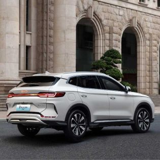 Rear-side view of a white BYD Song Plus DM-i SUV, showcasing its sleek design, full-width taillights, and stylish wheels, parked on a road in front of a grand stone building.