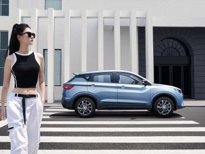 Woman (black crop top, white pants) walks by a light blue SUV on a crosswalk, modern building in the background.
