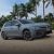 A gray Volkswagen Golf parked on sandy ground with palm trees in the background.