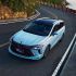 A light blue MPV drives on a coastal winding road, with lush greenery and the sea in the background, highlighting its sleek design and scenic appeal.