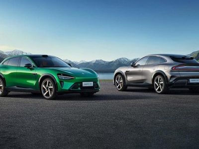 Two Xiaomi electric cars, one green and one silver, parked on a paved area with a scenic backdrop of mountains and a lake.