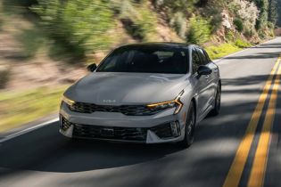 Sporty silver Kia K5 sedan driving on winding mountain road, showcasing aggressive front fascia and signature LED daytime running lights