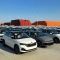 Numerous cars parked in an orderly manner at a port, with stacks of colorful containers in the background under a clear blue sky.