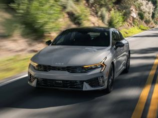 Sporty silver Kia K5 sedan driving on winding mountain road, showcasing aggressive front fascia and signature LED daytime running lights