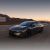 A dark gray Xpeng P7+ electric car driving on a road with mountains and a sunset sky in the background.