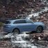 A light blue SUV driving through a stream with rocky terrain in the background.