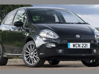 A black Fiat hatchback with license plate "WCN 221", parked on a gravel area with a rural landscape of trees and buildings in the background, showcasing its sleek design.