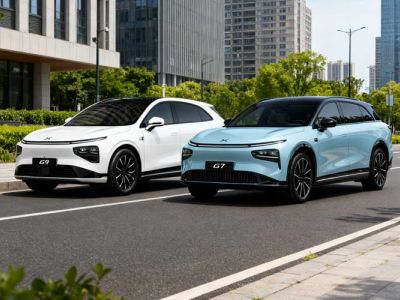 Two XPeng EVs (white G9, light blue G7) parked on a city street, modern buildings and greenery in the background.