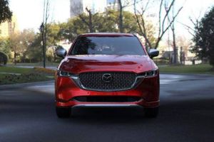 Front view of a red Mazda SUV, featuring its distinctive grille and Mazda logo, parked on a road in a park-like urban setting with trees and buildings in the background.