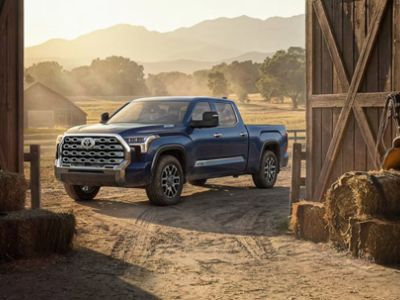 Three-quarter front view of blue Toyota crew cab pickup truck parked on rural farm dirt ground near wooden barn, rolling hills backdrop at golden hour