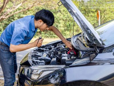 Auto repair technician leaning over a car with its hood open, inspecting and troubleshooting the vehicle engine outdoors in daylight