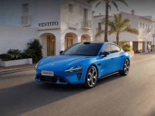 A bright blue Xiaomi SU7 sports sedan parked on a Mediterranean-style street with palm trees.