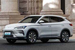 Side view of BYD Song Plus DM-i 2025 model, a light gray SUV with sleek design, parked on a road in front of a grand stone building, showcasing its elegant exterior.