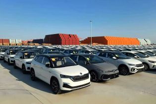 Numerous cars parked in an orderly manner at a port, with stacks of colorful containers in the background under a clear blue sky.