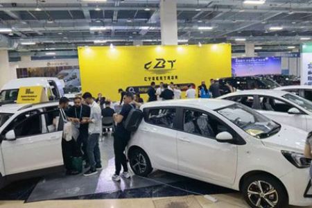 A car exhibition scene with several white cars on display, a large yellow "CBT CYBERTRON" backdrop, and people interacting, set in an indoor exhibition hall.